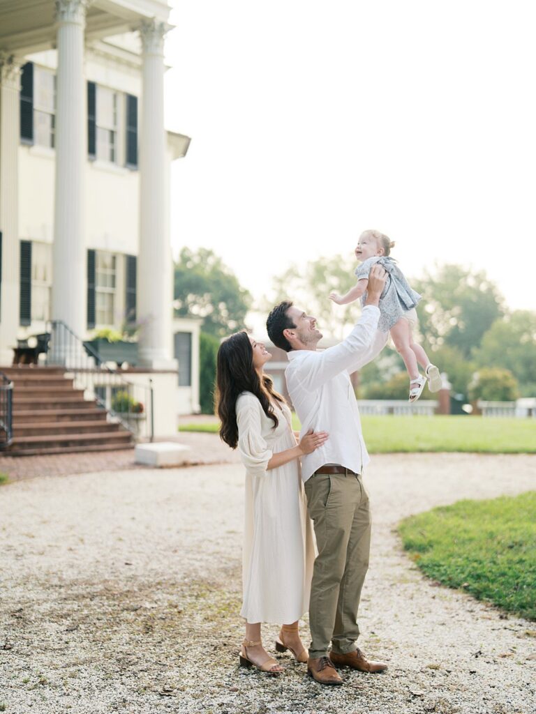 A Family Stands In Front Of A Grand Historic Home As A Father Lifts His Child Into The Air, Surrounded By Glowing Evening Light And Timeless Architecture.