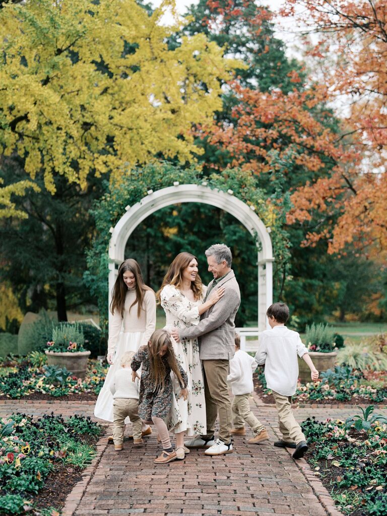 A Family Walks Together Through A Garden Arch Surrounded By Vibrant Fall Foliage, Capturing The Warmth And Connection That Makes You Wonder When Is The Best Time To Get Family Photos In The Dmv.