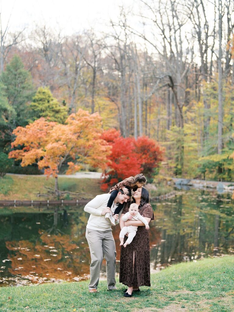 A Family Of Four Shares A Sweet Embrace By The Water With Vibrant Fall Foliage Behind Them, A Beautiful Example Of When Is The Best Time To Get Family Photos In The Dmv For Warm, Colorful Images.