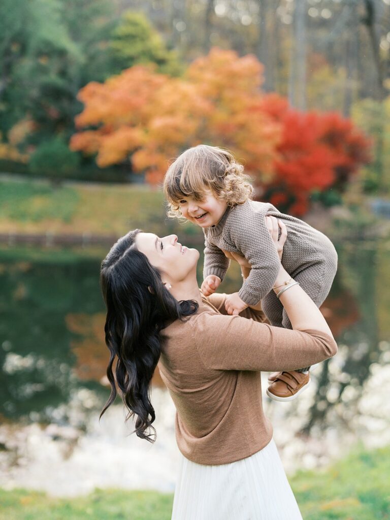A Mother Lifts Her Laughing Toddler By A Lakeside Surrounded By Rich Autumn Colors, A Joyful Moment That Reflects When Is The Best Time To Get Family Photos In The Dmv.