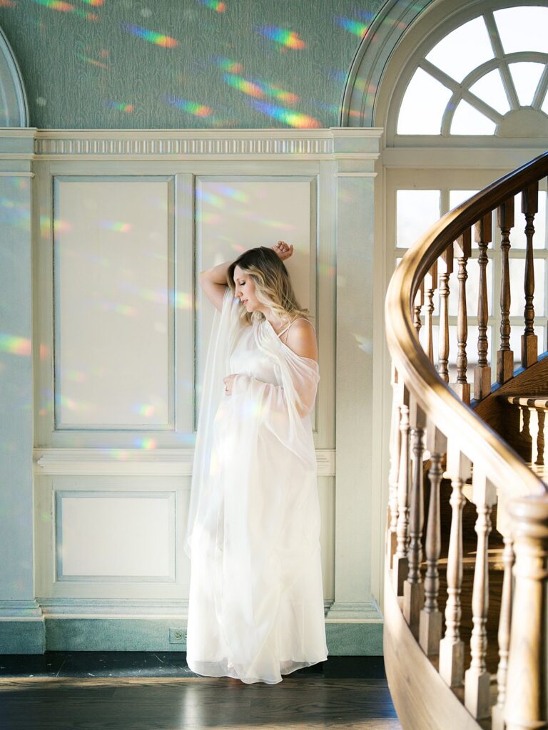 A Mother In A Flowing Gown Stands Near A Grand Staircase With Soft Rainbow Light Streaming Through The Windows, Capturing An Ethereal Indoor Moment That Reflects When Is The Best Time To Get Family Photos In The Dmv, Even Beyond Outdoor Seasons.