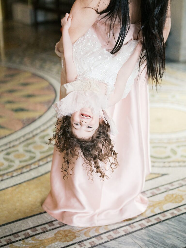 A Playful Moment As A Mother Lifts Her Child Upside Down, Capturing Joy And Movement Against Intricate Architectural Details.
