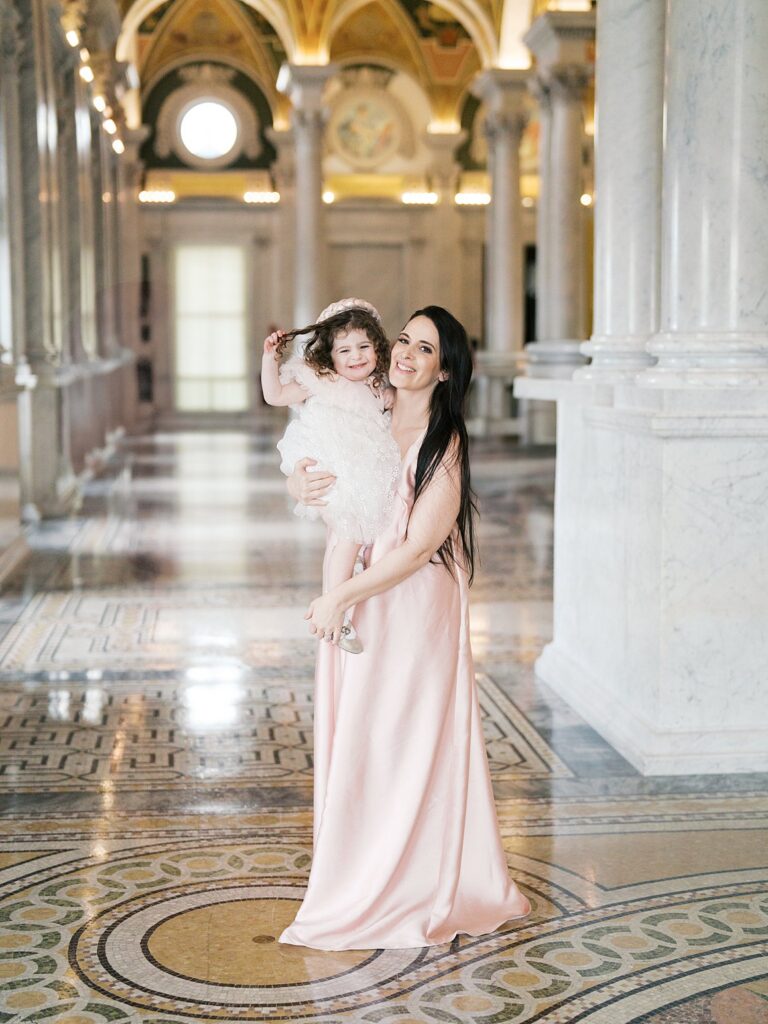 A Mother Holds Her Smiling Child In A Light-Filled Hallway With Marble Floors And Towering Columns, A Timeless Scene That Beautifully Shows When Is The Best Time To Get Family Photos In The Dmv Can Include Elegant Indoor Locations.