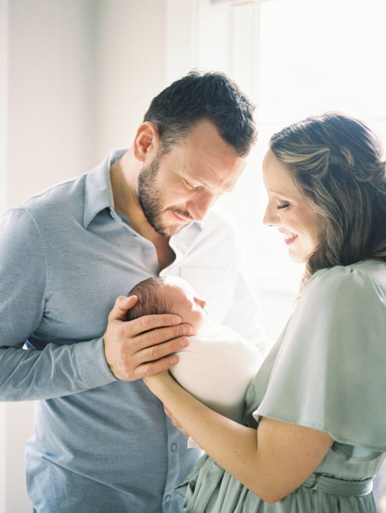Wondering How Long Does A Newborn Session Take? Long Enough To Capture A Glowing Moment Like This One, With Mom And Dad Together Cradling Their Newborn In Soft Window Light