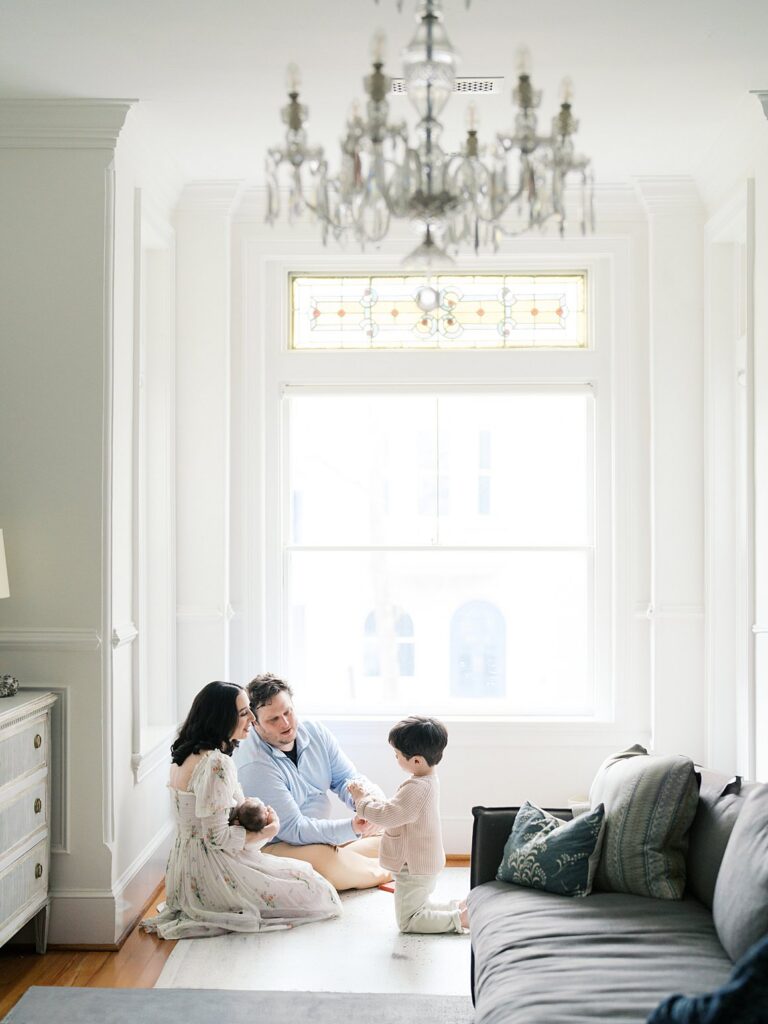 A Full Family Portrait Taken During A Newborn Session In Washington Dc, With Mom, Dad, And A Toddler Gathered Together On The Floor Of A Bright, Airy Room With Stained Glass Windows And A Chandelier Overhead