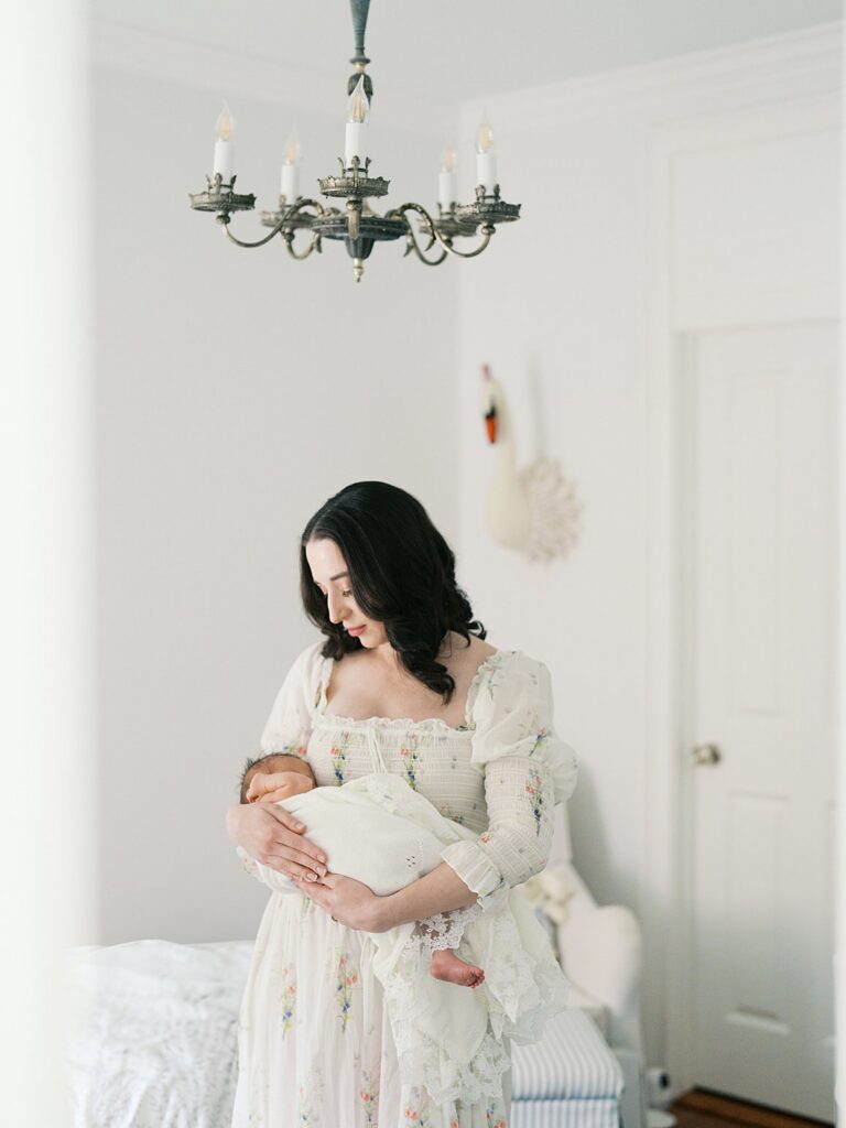 How Long Does A Newborn Session Take? Long Enough For Beautiful, Unhurried Moments Like This One, With A Dark-Haired Mama In A Dreamy Floral Dress Gazing Down At Her Newborn Beneath A Crystal Chandelier