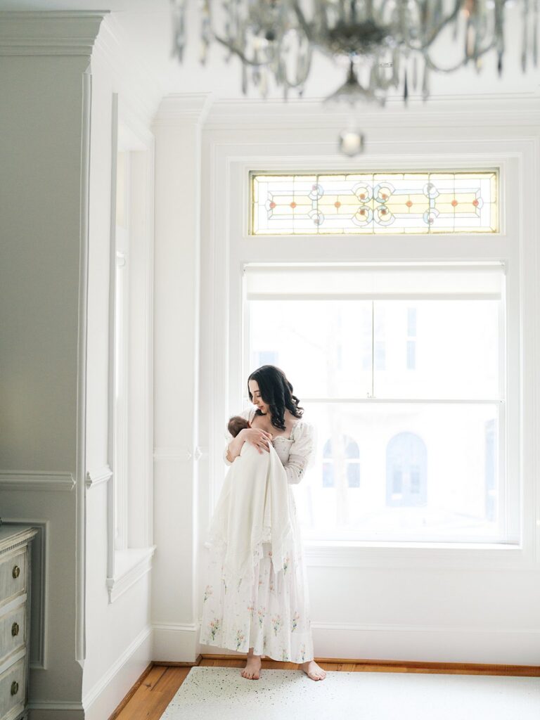 A Mother In A Flowing Floral Gown Holds Her Newborn By A Sunlit Stained Glass Window During A Newborn Session In Georgetown.