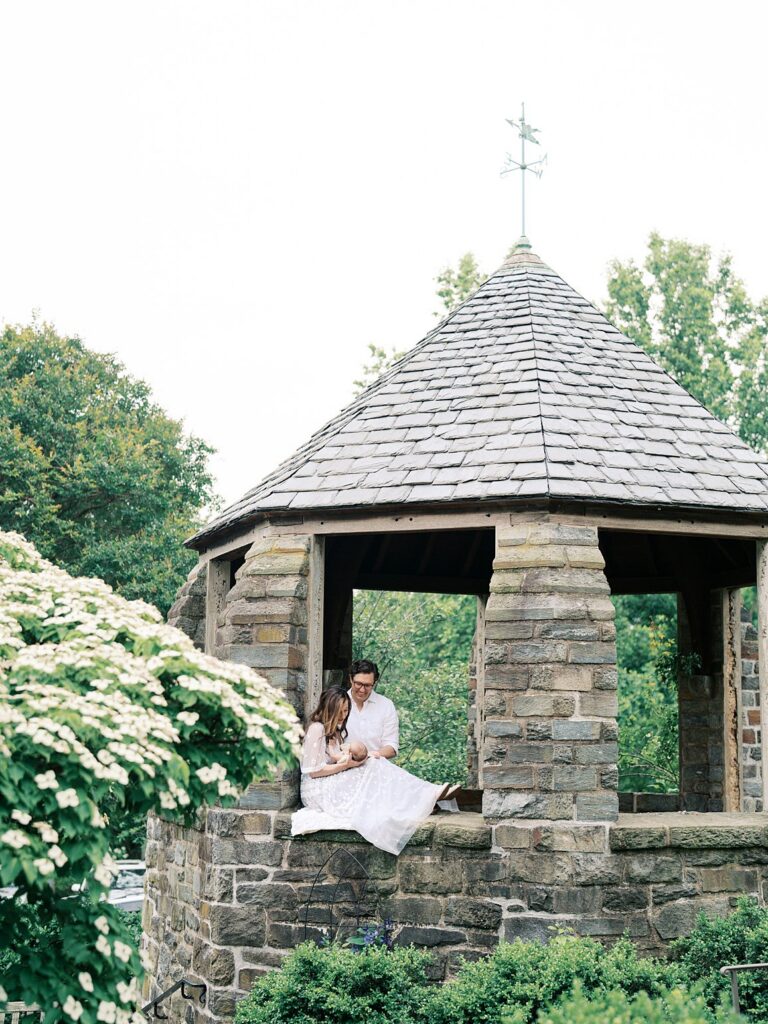 A Mother And Father Nestled Together In A Ivy-Covered Stone Gazebo Cradling Their Newborn, Surrounded By White Hydrangeas In A Dreamy Outdoor Newborn Portrait Session.