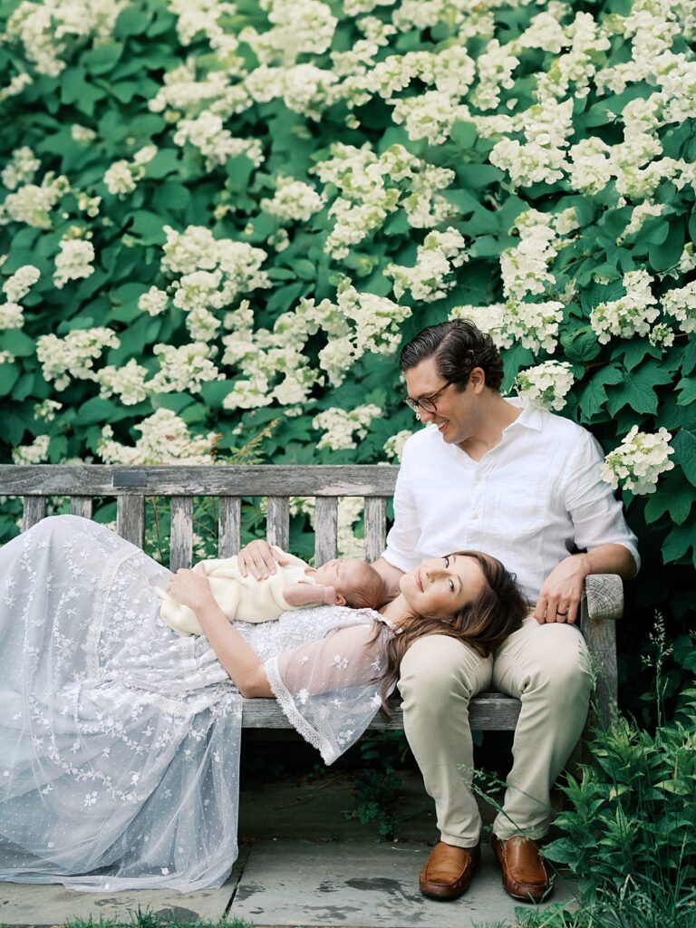 A Mother Reclines On A Garden Bench Holding Her Newborn While Her Husband Looks On Lovingly, Surrounded By Lush White Hydrangea Blooms During An Outdoor Fine Art Newborn Photography Session.