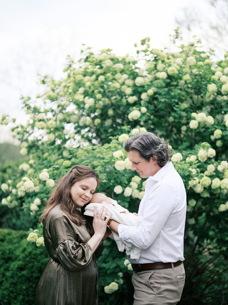 A Mother And Father Gazing Down At Their Sleeping Newborn Baby Surrounded By Lush White Hydrangeas During An Outdoor Newborn Photography Session.