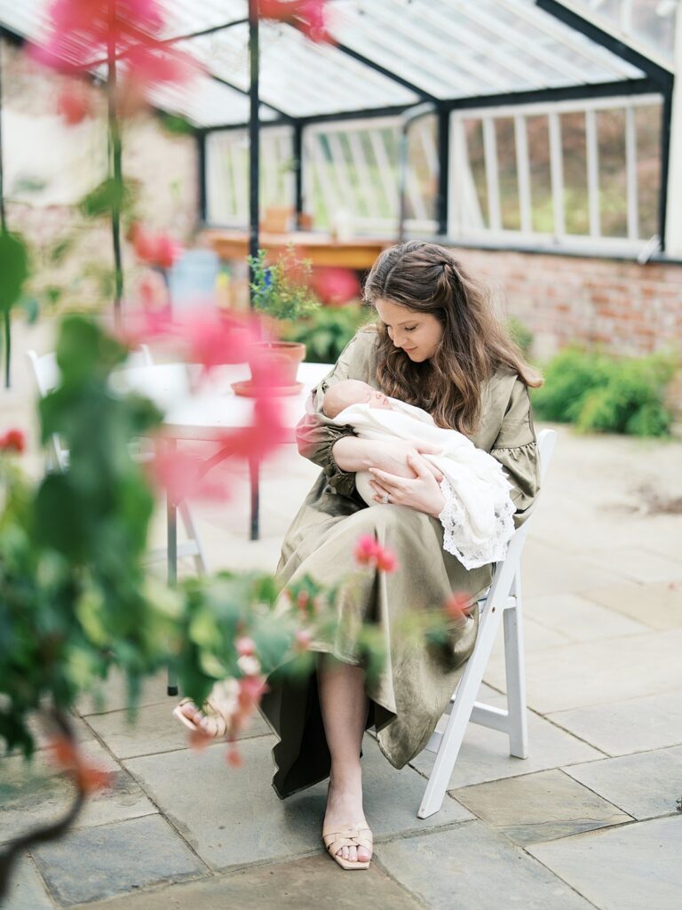 A Mother In A Sage Green Dress Seated In A Garden Greenhouse Setting, Cradling Her Newborn Baby During A Fine Art Outdoor Newborn Photography Session.