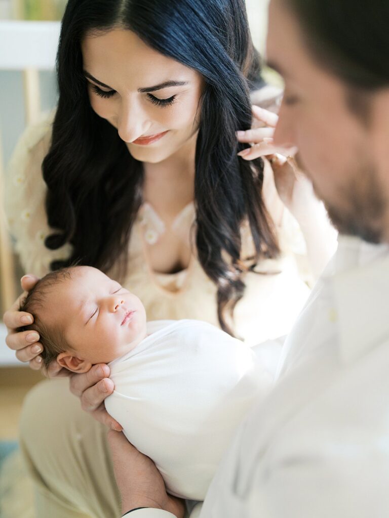 A Close-Up Of A Mother And Father Gazing Lovingly At Their Swaddled Sleeping Newborn, Capturing The Tender Early Days That Answer The Question Of How Old Should My Baby Be For Newborn Photos — This Peaceful Newborn Stage Is Exactly What We Aim To Photograph Within The First Two Weeks.