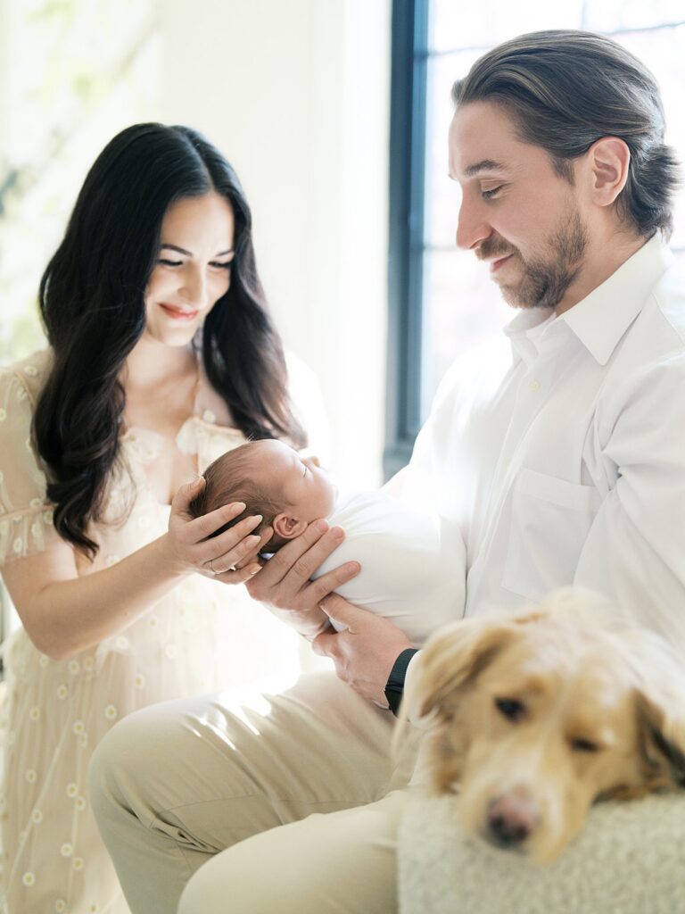 A Mother And Father Admiring Their Newborn Baby In Soft Natural Light During An In-Home Newborn Session, With The Family Dog Resting Nearby — A Beautiful Reminder That How Old Your Baby Is For Newborn Photos Affects How Peacefully They Sleep Through Sweet Moments Like This.
