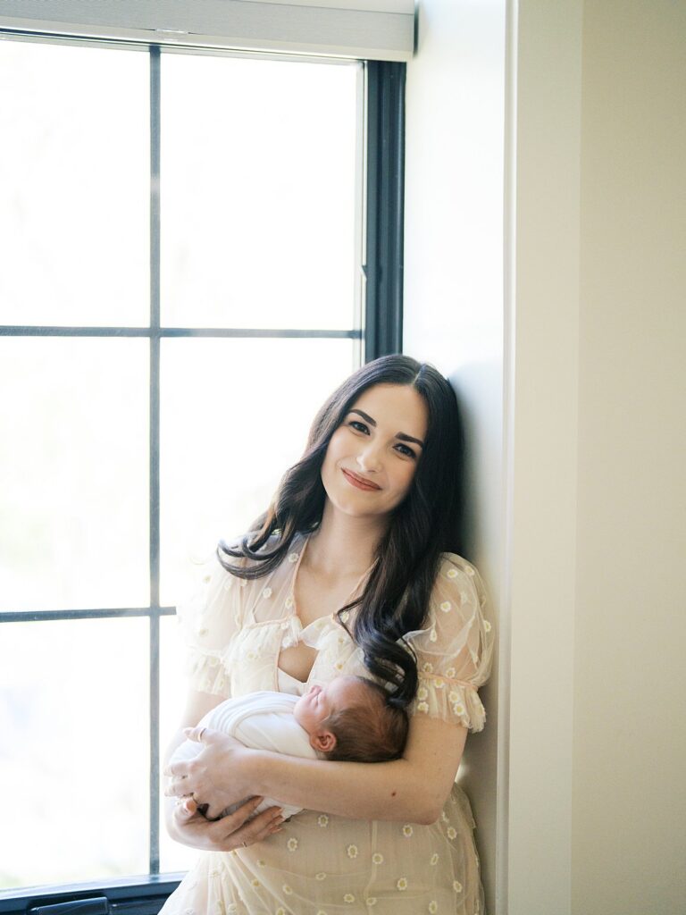 A Mother In A White Lace Dress Holding Her Newborn Baby By A Large Window In Soft Natural Light During An In-Home Fine Art Newborn Photography Session — A Gorgeous Example Of How Old Should My Baby Be For Newborn Photos Answered In Real Life, With A Sleepy Newborn In Those Precious First Days.