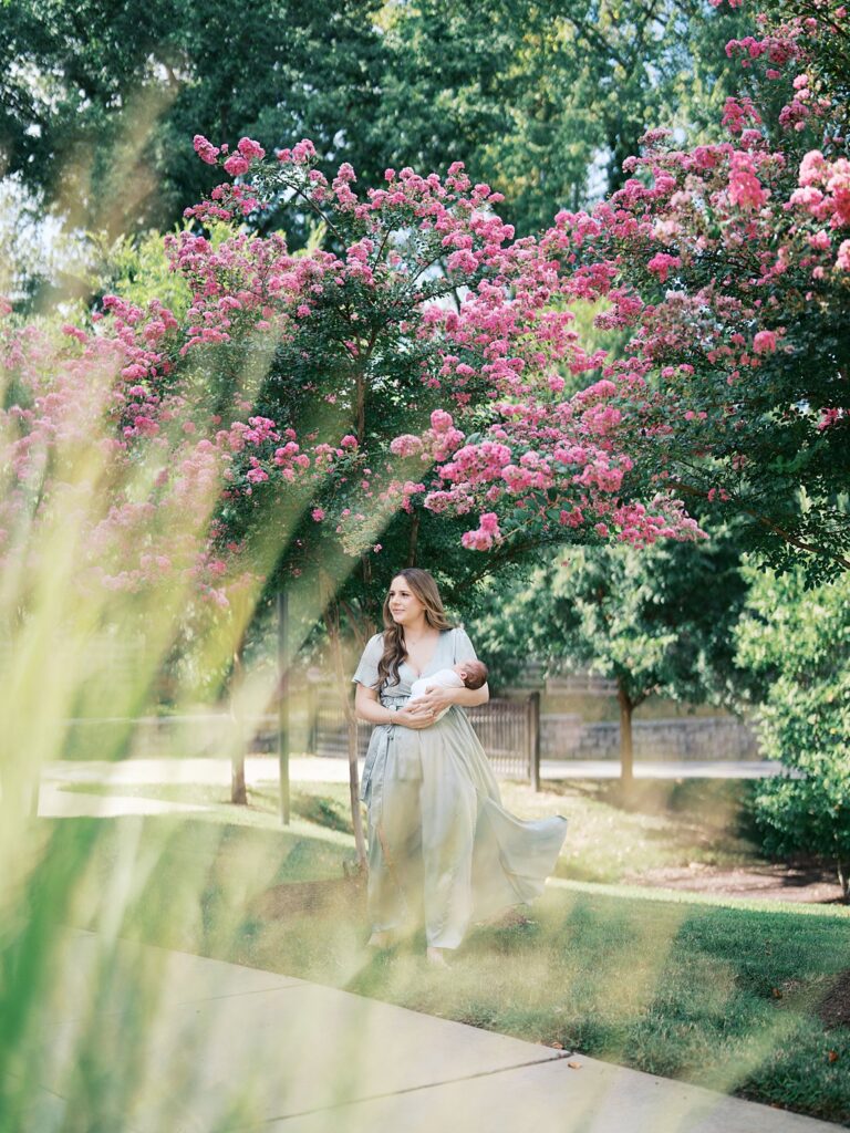 A Mother In A Flowing Sage Green Dress Walks Along A Garden Path Cradling Her Newborn, Framed By Towering Pink Crepe Myrtle Trees In Full Summer Bloom — For Every Parent Wondering &Quot;What If I Missed The Newborn Window For Photos,&Quot; This Is Proof The Magic Is Still There.