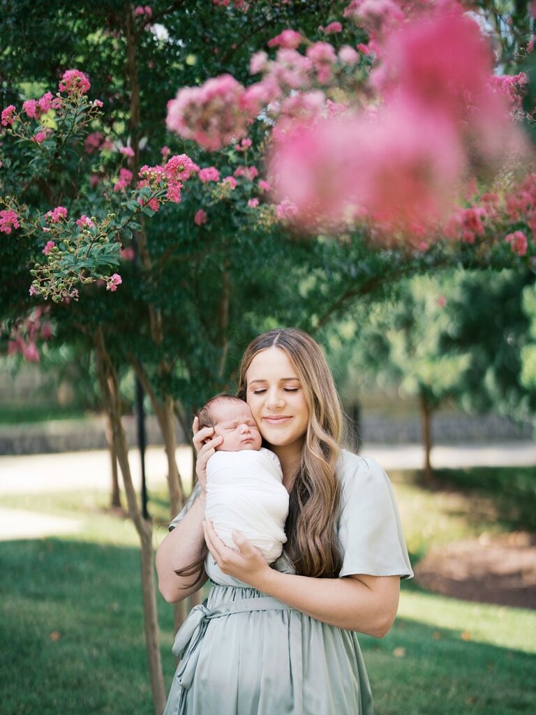 A Mother In A Sage Green Dress Closes Her Eyes And Smiles As She Cuddles Her Swaddled Newborn Against Her Cheek Beneath A Blooming Pink Crepe Myrtle Tree During An Outdoor Newborn Session.
