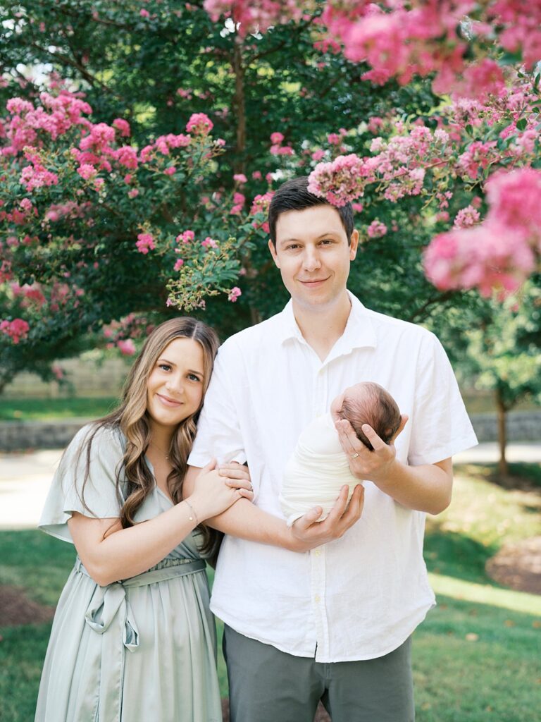 A Mother And Father Stand Together Beneath Blooming Pink Crepe Myrtle Trees, She Leaning Into Him While He Cradles Their Swaddled Newborn, Both Smiling Warmly At The Camera During An Outdoor Summer Newborn Session.