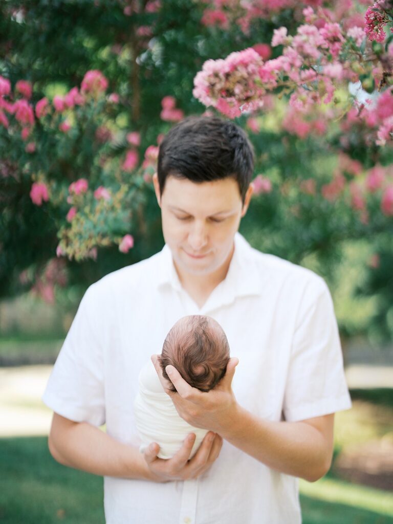 A Father In A White Button-Down Shirt Gazes Lovingly Down At His Swaddled Newborn Cradled In His Arms, With Soft Pink Crepe Myrtle Blossoms Blurred In The Background — Showing That It's Never Too Late To Ask &Quot;What If I Missed The Newborn Window For Photos&Quot; And Book A Session Anyway.
