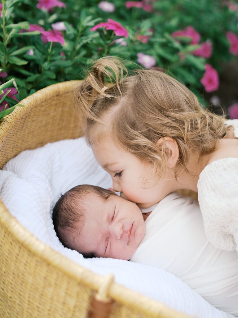 A Young Toddler Girl With Blonde Hair Gently Kisses Her Sleeping Newborn Sibling Nestled In A Wicker Bassinet Surrounded By Pink Garden Flowers — Captured During An Outdoor Newborn Session Before The Newborn Window For Photos Closes.