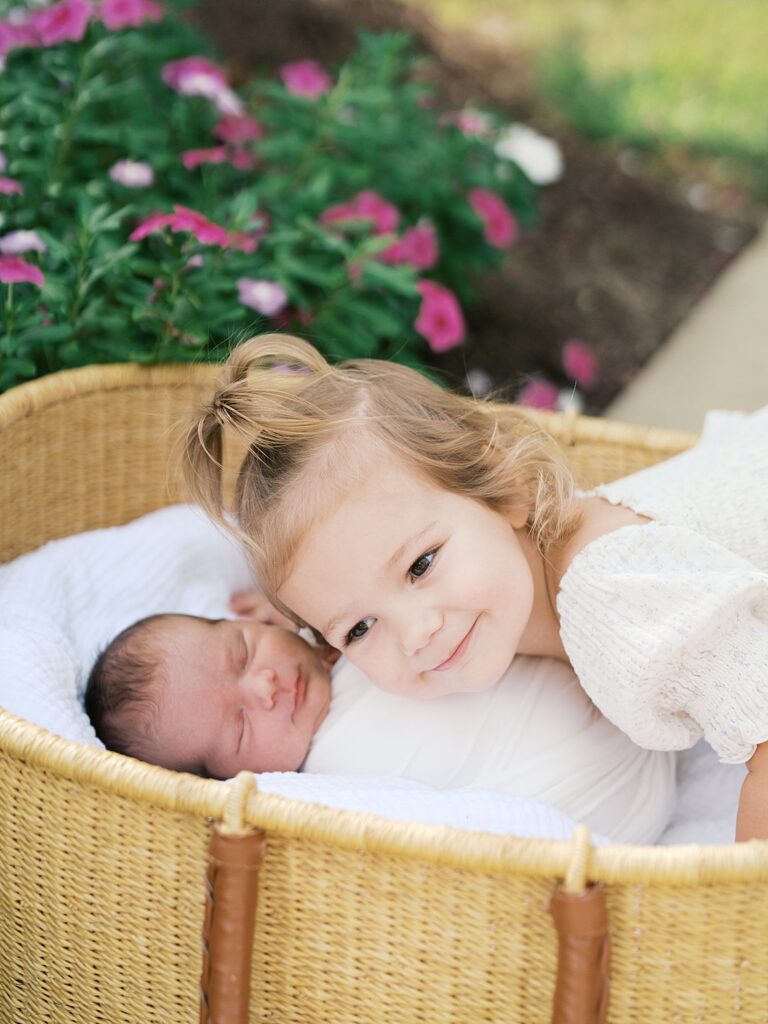 A Smiling Toddler Girl In A White Top Rests Her Chin On The Edge Of A Wicker Bassinet, Cheek To Cheek With Her Sleeping Newborn Sibling, Surrounded By Pink Garden Blooms — The Sweetest Sibling Moment From An Outdoor Newborn Session.