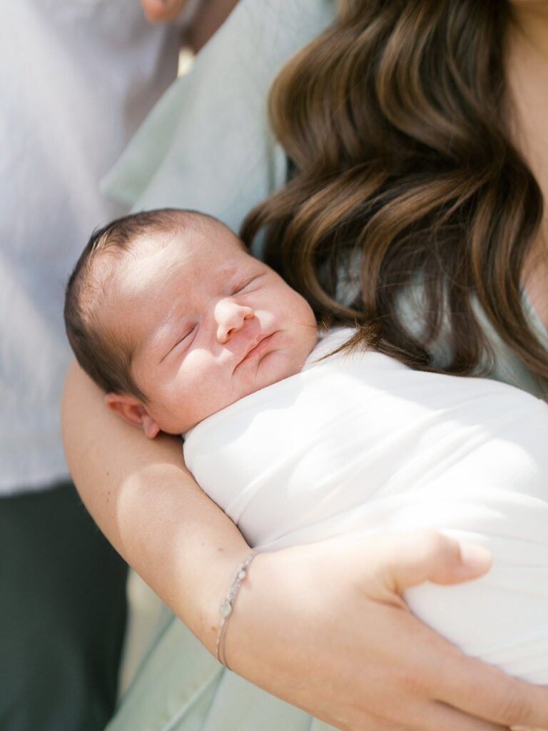 A Close-Up Of A Swaddled Newborn Sleeping Peacefully Against A Mother's Chest, Her Dark Wavy Hair Falling Softly Around Them — An Intimate Detail Shot From An Outdoor Newborn Session Capturing How Tiny And Fleeting This Season Truly Is.