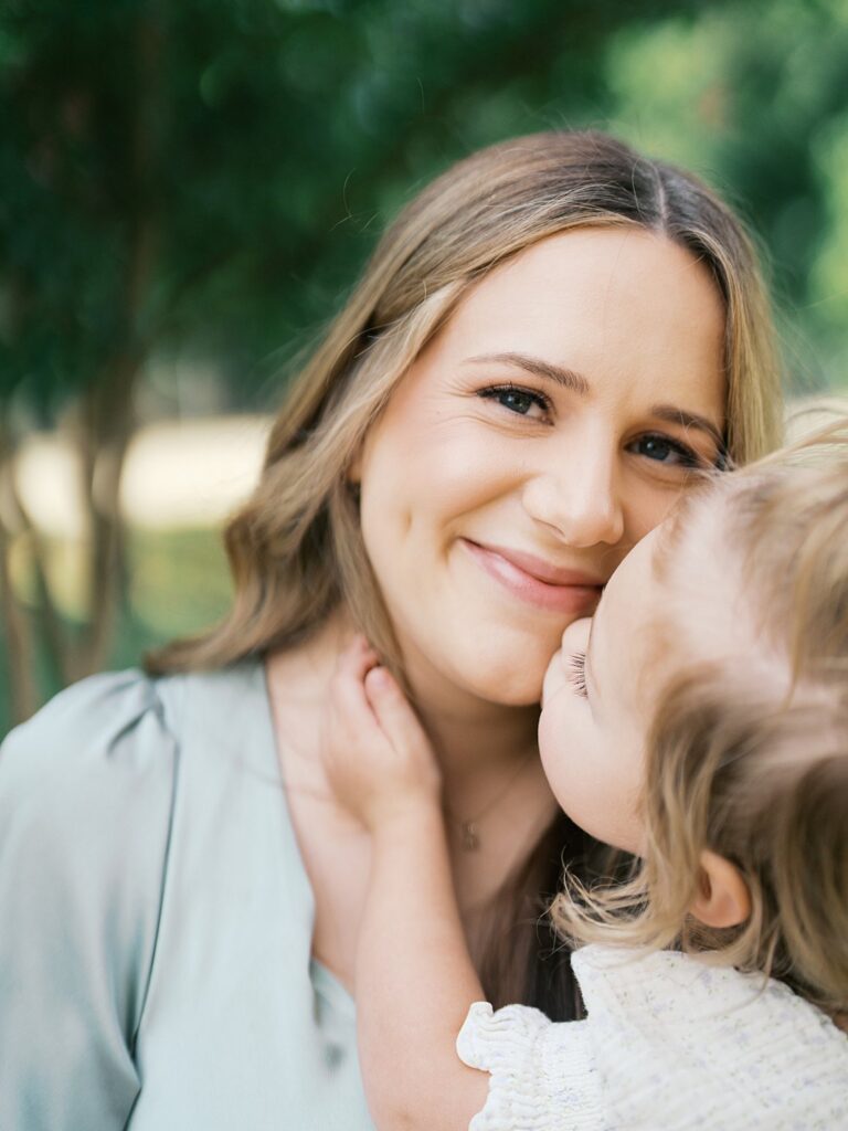 A Close-Up Portrait Of A Smiling Mother As Her Toddler Daughter Leans In To Kiss Her Cheek, Both Bathed In Soft Natural Light During An Outdoor Newborn Session — The Kind Of In-Between Moment That Makes A Lifestyle Session So Worth It.