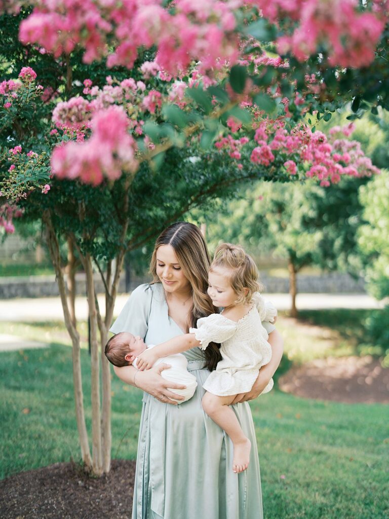 A Mother In A Sage Green Dress Holds Both Her Sleeping Newborn And Her Toddler Daughter Beneath A Blooming Pink Crepe Myrtle Tree — Proof That Even If You Missed The Newborn Window For Photos, A Lifestyle Newborn Session Can Still Beautifully Capture This Fleeting Season.