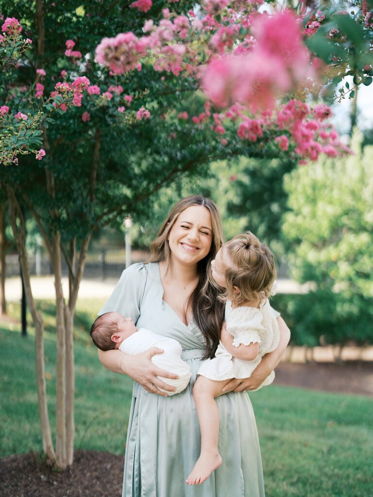 A Laughing Mother In A Sage Green Dress Holds Both Her Newborn And Her Toddler Daughter Beneath A Blooming Pink Crepe Myrtle Tree, Her Joy Completely Unposed — For Any Parent Who Has Wondered &Quot;What If I Missed The Newborn Window For Photos,&Quot; This Moment Is Your Answer.