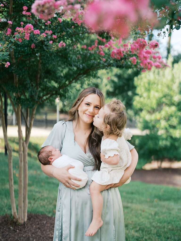 A Smiling Mother In A Sage Green Dress Holds Her Newborn While Her Toddler Daughter Leans In To Give Her A Kiss On The Cheek, Beneath A Blooming Pink Crepe Myrtle Tree During An Outdoor Newborn Session.