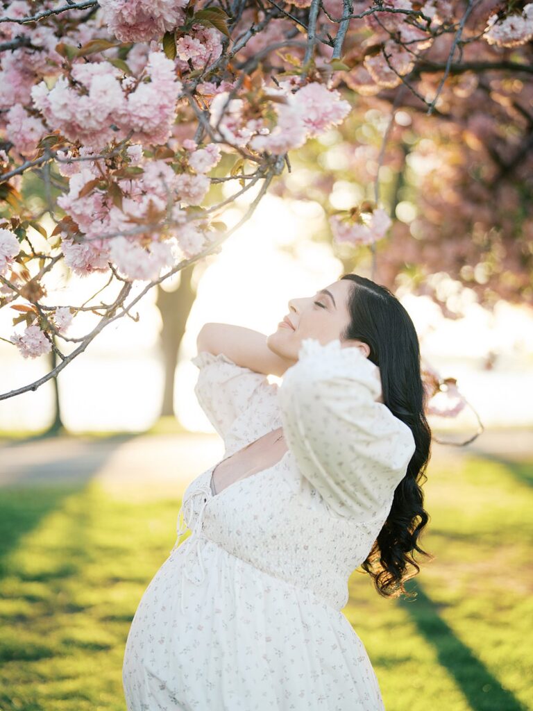 Maternity Portrait Under Pink Cherry Blossoms At Sunset With Glowing Backlight And Soft Pastel Tones, Capturing A Romantic And Whimsical Spring Motherhood Session.