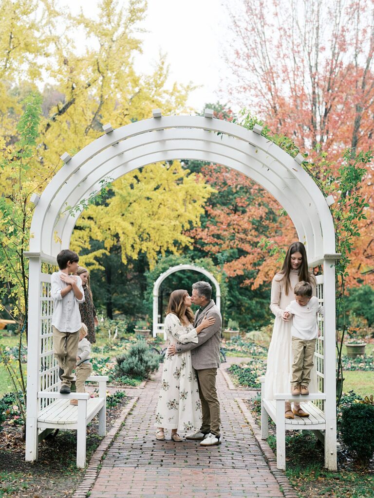 Family Of Five Gathered Beneath A White Garden Arbor With Vibrant Yellow And Red Fall Leaves Creating A Whimsical And Colorful Outdoor Portrait Backdrop.