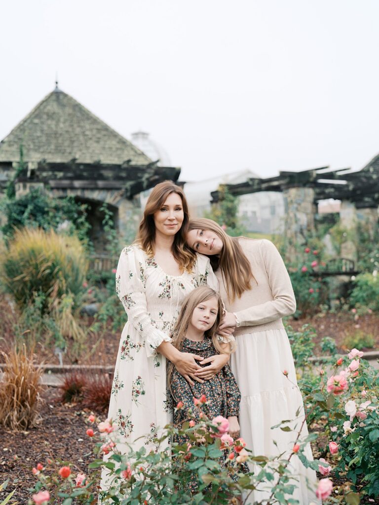 Mother And Daughters Posing Among Blooming Roses And Soft Autumn Greenery During A Romantic Family Photography Session At A Historic Garden.