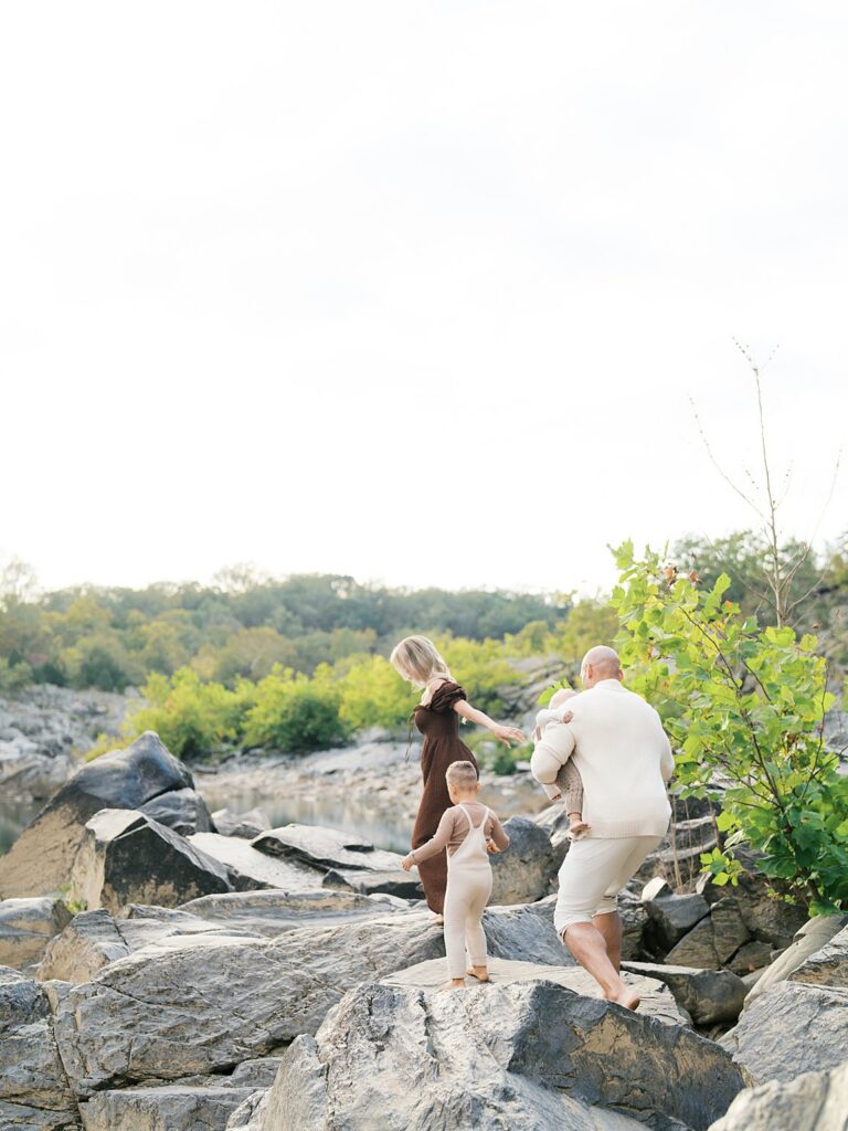 Parents Walking Hand-In-Hand With Their Young Child Across Rocky Terrain During A Light-Filled Outdoor Family Session With Earthy Neutral Tones And A Candid Storytelling Feel.