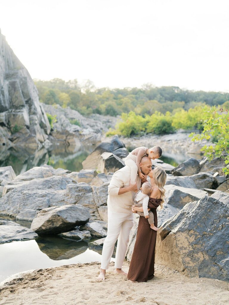 Family Of Three Sharing A Quiet Moment Among Rocky Cliffs And Soft Water Reflections During An Adventurous Outdoor Family Photography Session At Sunset At Great Falls In Maryland.