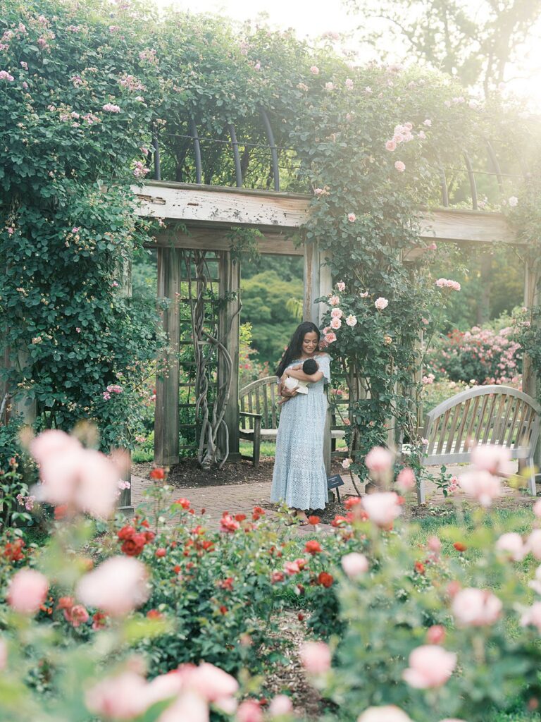 Mother Holding Her Newborn Baby Beneath A Rose-Covered Garden Arbor During A Romantic Outdoor Newborn Photography Session Filled With Soft Summer Blooms.