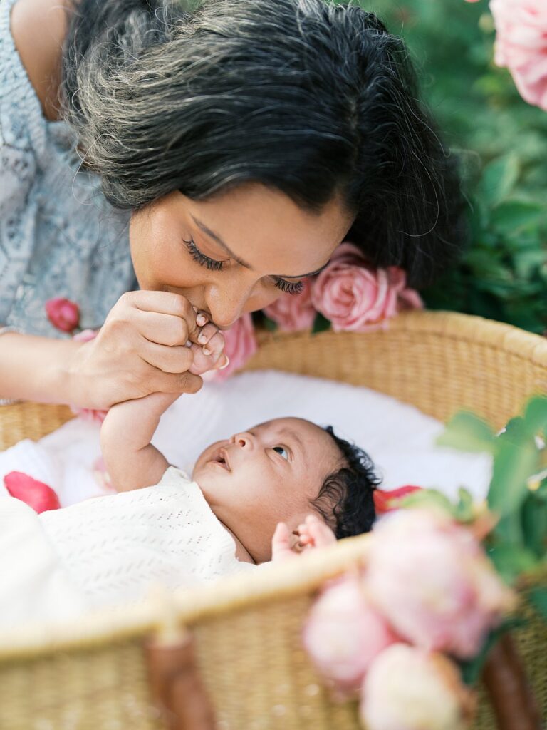 Close-Up Of A Mother Kissing Her Newborn Baby’s Tiny Hand While The Baby Rests In A Wicker Bassinet Surrounded By Pastel Roses And Greenery.