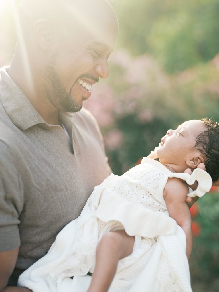 Father Smiling Down At His Newborn Baby During A Sun-Drenched Outdoor Newborn Photography Session With Warm Golden Light And Soft Spring Blooms In The Background.