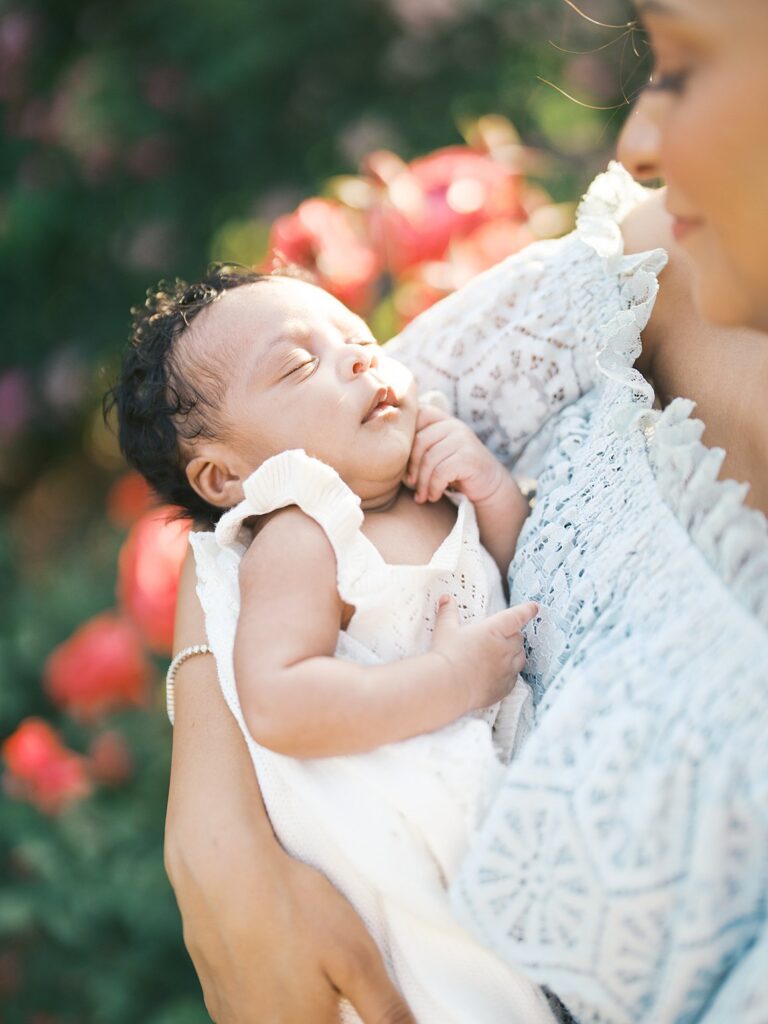 Close-Up Newborn Portrait Of A Sleeping Baby Girl Wrapped In Her Mother’s Arms During A Dreamy Outdoor Newborn Session With Soft Floral Tones And Glowing Evening Light.