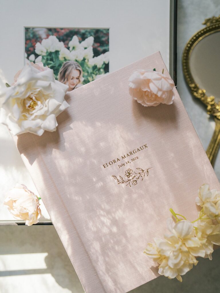 Fine Art Heirloom Album With Gold Embossed Lettering Displayed In Natural Window Light With Delicate Flowers, Celebrating Tangible Printed Memories In The Digital Age.