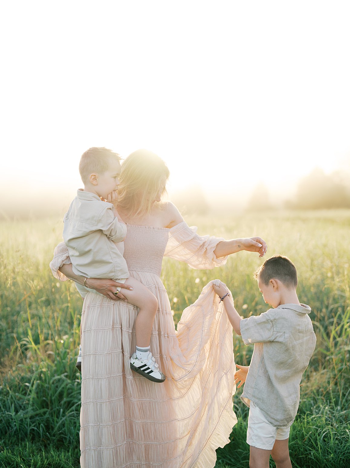 Mother In A Blush Dress Holding Her Young Son In Tall Golden Grass While Another Boy Holds Her Dress Hem, Photographed By A Gainesville Photographer