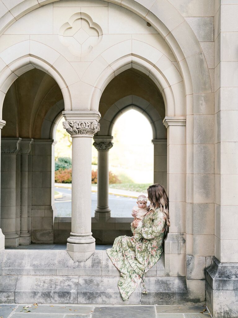 Mother Holds Her Baby Beneath The Stone Gothic Arches Of The Bishop's Garden, One Of The Most Magical Washington Dc Fall Foliage Locations For A Family Session.