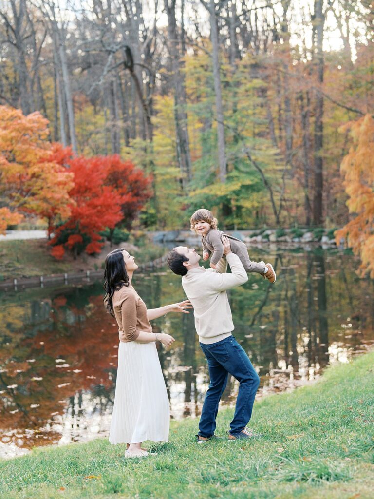 Parents Lift Their Toddler Beside A Pond Framed By Red And Gold Trees At Brookside Gardens, One Of The Best Washington Dc Fall Foliage Locations For A Family Session.