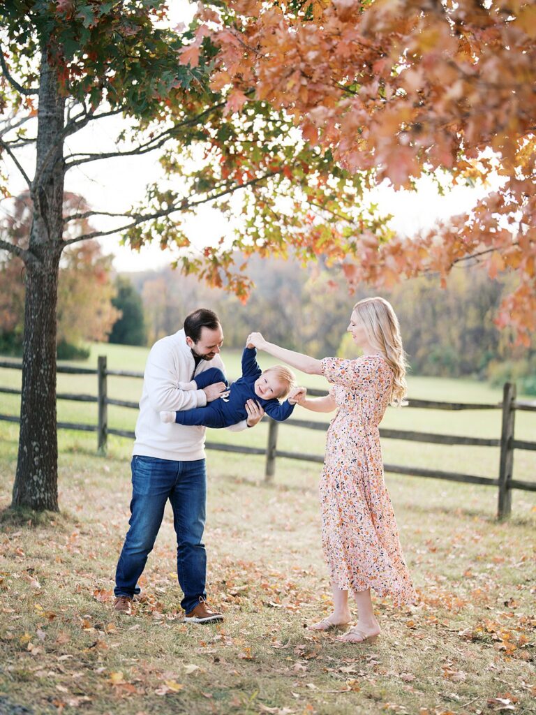 Parents Play With Their Toddler Under A Golden Autumn Oak By A Split-Rail Fence, A Fall Family Session At One Of The Best Washington Dc Fall Foliage Locations.