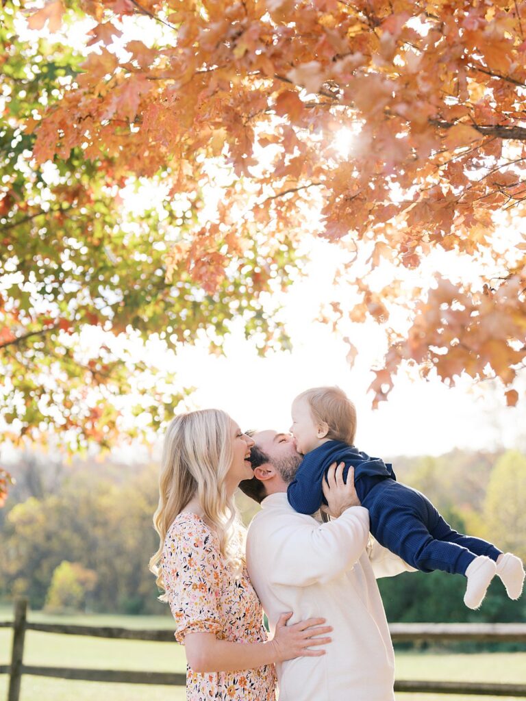 Parents Kiss And Lift Their Little One Under A Glowing Golden Oak At Oatlands, A Favorite Among Washington Dc Fall Foliage Locations.