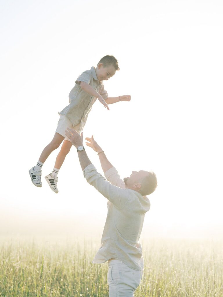 Father Tossing His Young Son Into The Air Against A Bright Sunrise Sky During A Golden Hour Family Session With A Gainesville Photographer