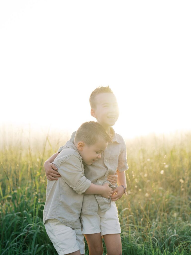 Two Young Brothers Hugging In Tall Golden Grass At Sunrise During A Manassas Battlefield Family Session