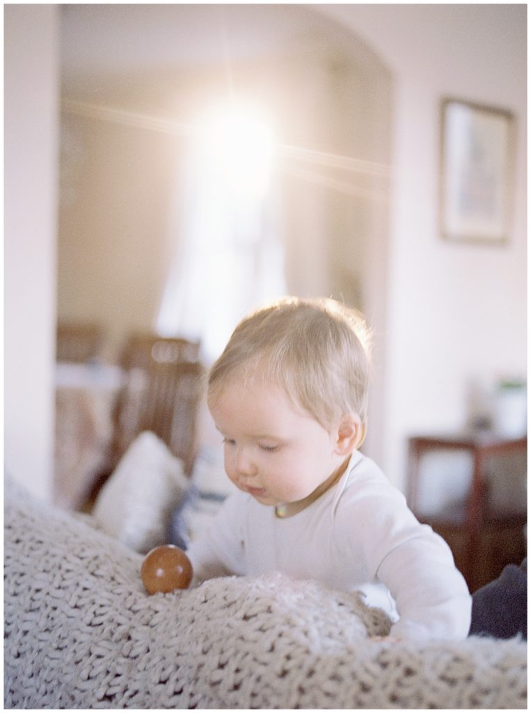 A baby standing up on the couch while the light shines through the window.