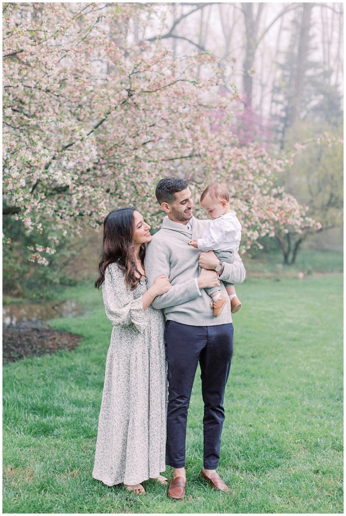 Mother, baby, and father in front of a cherry blossom in Brookside Gardens during a family photo session.