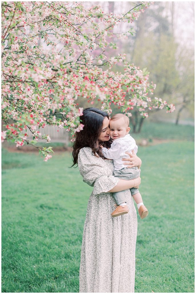 Mother snuggles with her baby in front of a cherry blossom tree at Brookside Gardens