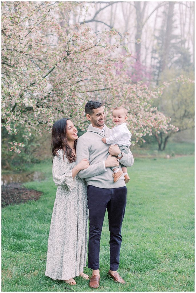 Mother, father, and baby boy stand by a cherry blossom tree in Brookside Gardens in Maryland